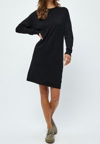 Una donna sta in piedi con sicurezza indossando un vestito nero a maniche lunghe e scarpe slip-on verde oliva, su uno sfondo chiaro.