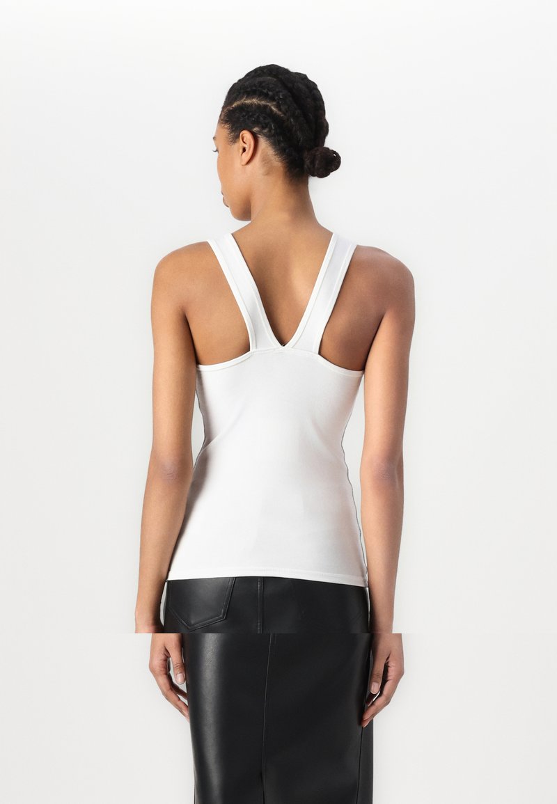 Top blanc sans manches avec des bretelles larges en V, fabriqué dans un tissu lisse et extensible, offrant une silhouette ajustée.