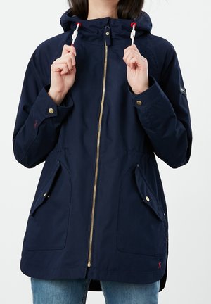 Persona con chaqueta con capucha azul marino con cremallera dorada y grandes bolsillos delanteros, sosteniendo los cordones blancos cerca de la capucha.