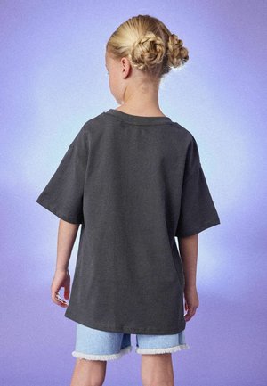 Pelēka oversize kokvilnas t-krekls kopā ar gaišiem džinsa šortiem ar saplēstām malām. Fonā ir maigs violetas krāsas gradients.