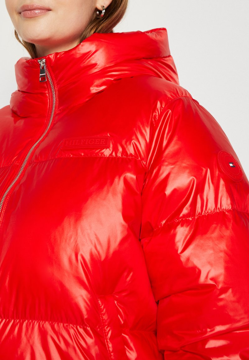 Giacca puffer rossa con colletto alto, finitura lucida, chiusura con zip e dettaglio del marchio sul petto e sulle maniche. Texture morbida e imbottita.