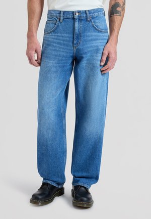 Άνδρας που φοράει φαρδιά μπλε τζιν και μαύρα δερμάτινα παπούτσια, στέκεται μπροστά σε απλό φόντο, με εμφανές τατουάζ στο χέρι.