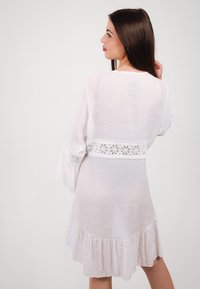 Vestito bianco leggero con maniche lunghe, bordo in pizzo in vita e orlo arricciato. Texture liscia e design fluido.