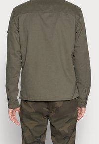Uomo che indossa una camicia a maniche lunghe verde oliva e pantaloni verde mimetico, visto di spalle su uno sfondo neutro.