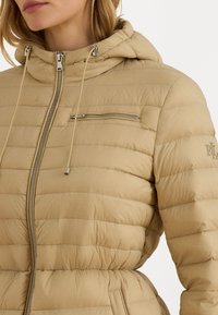 Jachetă puffer bej cu cusături orizontale, o buzunar cu fermoar pe piept și o glugă cu șnururi ajustabile. Textură moale și netedă.