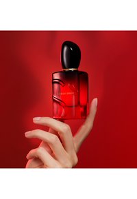 Main tenant un flacon de parfum Giorgio Armani rouge et noir sur fond rouge.