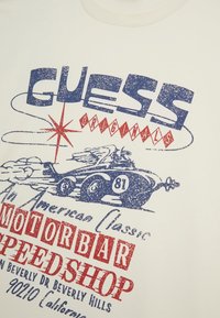 T-shirt bianca in cotone con stampa grafica blu e rossa di un'auto d'epoca con testo "GUESS Originals" e "Motorbar Speedshop".