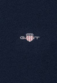 Tissu en coton bleu marine avec un logo de bouclier orné d'étoiles et de rayures, avec le texte blanc "GANT" centré sous l'emblème.