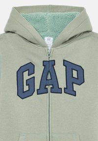 Zelená mikina na zips s mäkk�ým mätovozeleným interiérom. Obsahuje veľké námornícke písmená "GAP" na prednej strane a kapucňu.