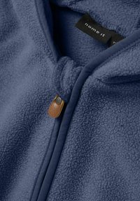 Primer plano de una tela de forro polar azul con bordes cosidos, una pequeña pestaña de cuero marrón, y una etiqueta de ropa negra parcialmente visible.