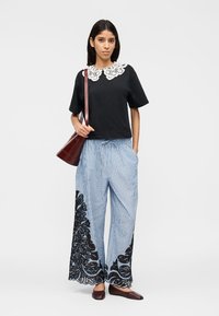 Femme portant un haut noir avec un col en dentelle blanche, un pantalon large à rayures bleues avec des motifs en dentelle noire, des chaussures plates marron, et portant un sac à bandoulière marron.