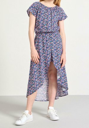 Ung person iført en blå blomsterkjole med korte ærmer og en frontslids, kombineret med hvide sneakers, stående på en lys baggrund.