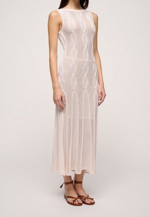 Jasnoróżowa, dzianinowa sukienka maxi bez rękawów, z ażurowymi wzorami i dopasowanym krojem. Noszona z brązowymi sandałami na paseczki.
