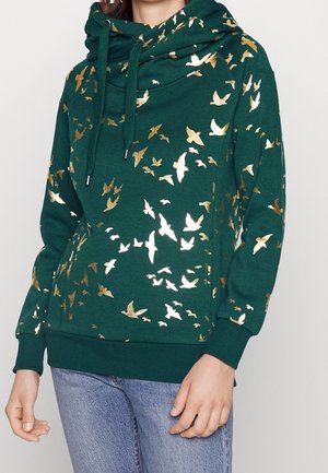 Kobieta w ciemnozielonej bluzie z kapturem z złotymi wzorami ptaków i niebieskich dżinsach, stojąca z rękami swobodnie opuszczonymi wzdłuż ciała.