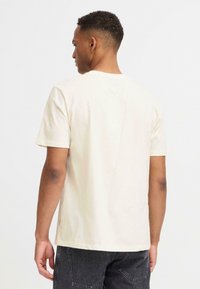 Lys beige bomulls t-skjorte med korte ermer og rund hals, vist fra baksiden, med en avslappet passform og minimalt design.