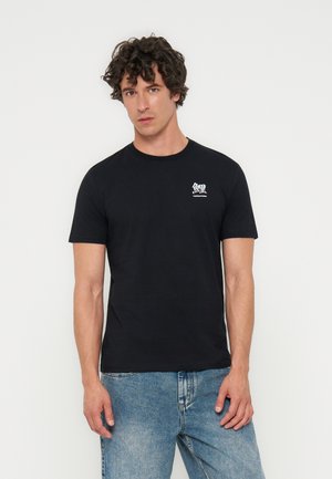 Giovane uomo con capelli ricci che indossa una maglietta nera con testo giapponese bianco e jeans azzurri, in piedi davanti a uno sfondo bianco semplice.