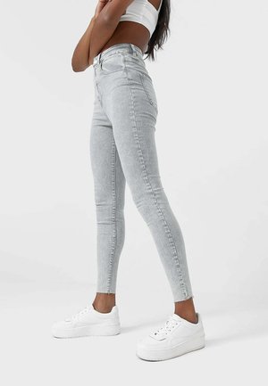 Femme portant un jean skinny gris clair et des baskets blanches, debout, bras croisés, sur un fond uni.