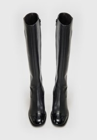 Une paire de bottes noires en cuir, hautes, avec des orteils carrés brillants et des fermetures éclair sur les côtés, se tenant debout sur un fond blanc uni.