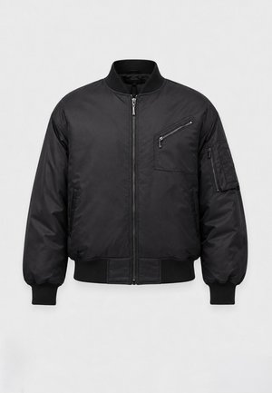 Homme portant une veste bomber noire, un pantalon beige ample et des chaussures noires, debout devant un fond clair uni.