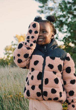 Niño sonriente al aire libre con una chaqueta peluda rosa con manchas negras, cubriéndose un ojo con la mano enguantada en una zona de césped con árboles.