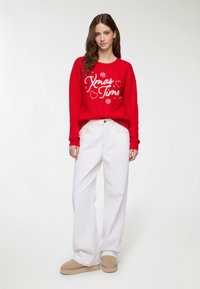 Czerwony sweter dzianinowy z napisem "Czas na święta" i sercami. Połączony z białymi spodniami w szerokim kroju i beżowymi kapciami. Gładka faktura i luźny krój.