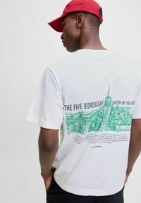 White cotton t-shirt with green graphic illustration of buildings and text on back, crew neck, short sleeves, worn with a red cap.