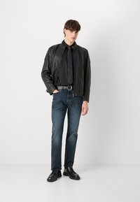 Black leather jacket with shoulder patches, black shirt and tie, blue denim jeans, and black shoes with a chunky sole in a neutral background.