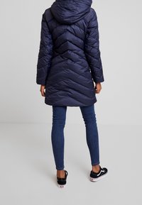 Marinblå puffjacka med chevron-sömnad, huva och långa ärmar. Tillsammans med mörka denimjeans och svarta sneakers. Slät, glänsande yta.