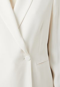 Blazer ajustado color blanco roto con solapa muestrade, cierre de un solo botón y un bolsillo lateral con un sutil detalle a rayas. Textura de tejido suave.