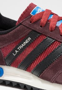 Röda och svarta idrottsskor med en överdel av mocka och texturerat nät, med tre svarta ränder och en blå tunganmäla. Vit mellansula.