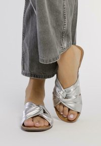 Sandalias de slide metálicas plateadas con un diseño anudado. Textura suave, punta abierta y suela marrón. Combinadas con pantalones grises de pierna ancha.