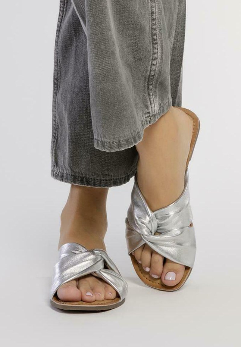 Sandalias de slide metálicas plateadas con un diseño anudado. Textura suave, punta abierta y suela marrón. Combinadas con pantalones grises de pierna ancha.