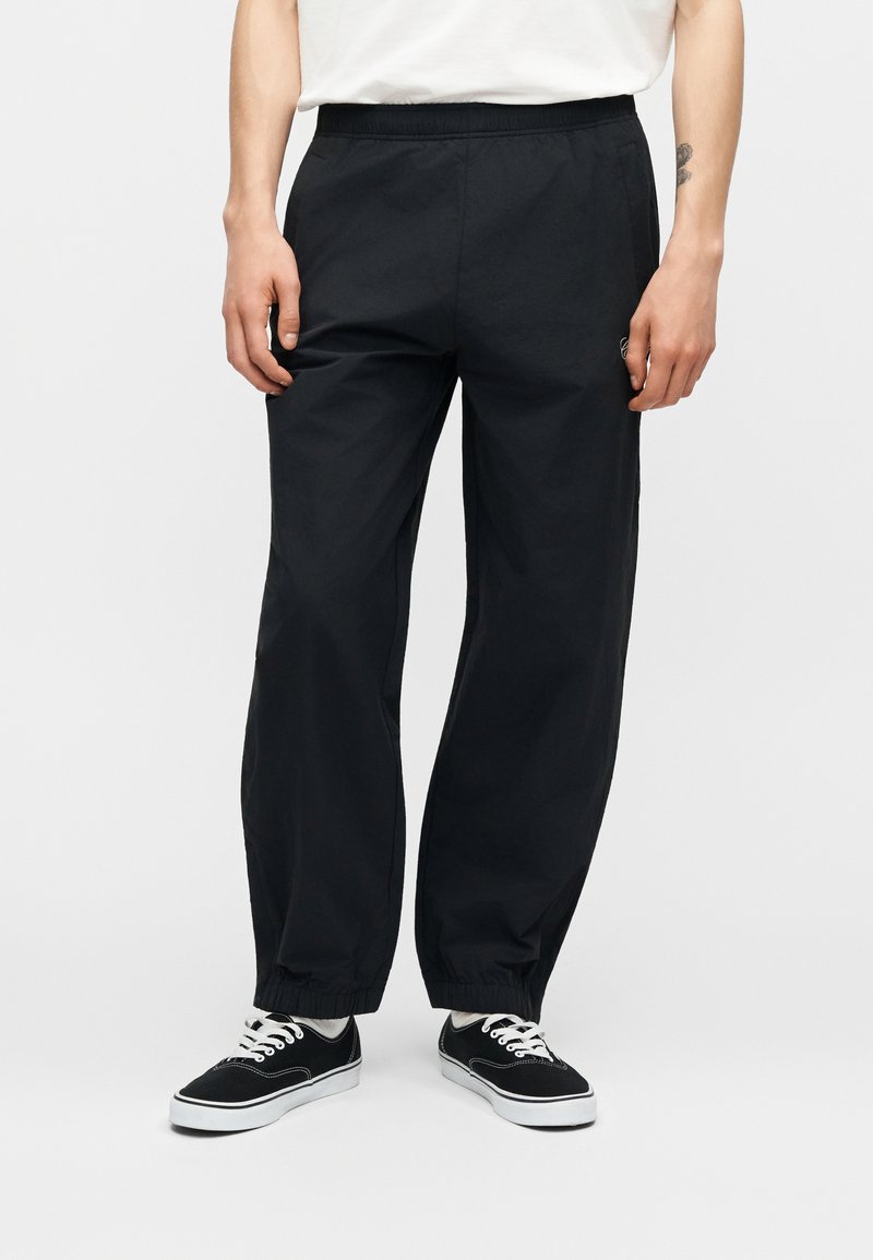 Person trägt schwarze Hosen mit elastischem Saum, weiße Socken, schwarze Schnürschuhe und ein weißes T-Shirt und steht vor einem weißen Hintergrund.
