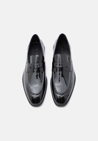 Fekete lakkbőr mokaszinok sima felülettel, lekerekített orral és széles pánttal az elülső részen. Lapos talp, minimális varrás.