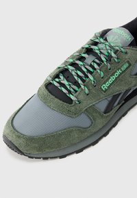 Πράσινο σνίκερ  από σουέντ και δίχτυ με μαύρες λεπτομέρειες, διαθέτει υφή κορδονιών και λαστιχένια σόλα. Το λογότυπο της Reebok εμφανίζεται ευδιάκριτα.