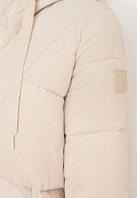 Nærbillede af en beige fløjlspufferjakkeærm med et præget logo-patch og matchende snoretræk med tekst.