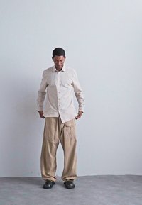 Man som bär beige oversized byxor, ljus rutig långärmad skjorta och svarta skor står mot en enkel ljusgrå vägg.