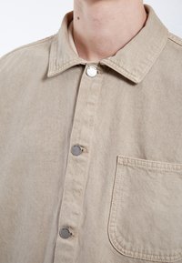 Camisa de mezclilla beige con cuello puntiagudo, bolsillo frontal y cierre de botón plateado. Textura suave con detalles de costura visibles.