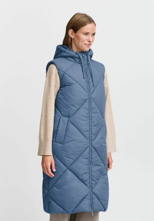 Gilet rembourré bleu avec capuche, présentant un motif matelassé en losanges, fermeture éclair à l'avant et poches latérales. Porté par-dessus un haut beige à manches longues.