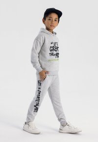 Grå bomullshoodie med grafiken "Låt oss skapa konsten" i svart och grönt. Matchande gråa sweatpants med sidofickor och liknande textdesign. Vita sneakers.