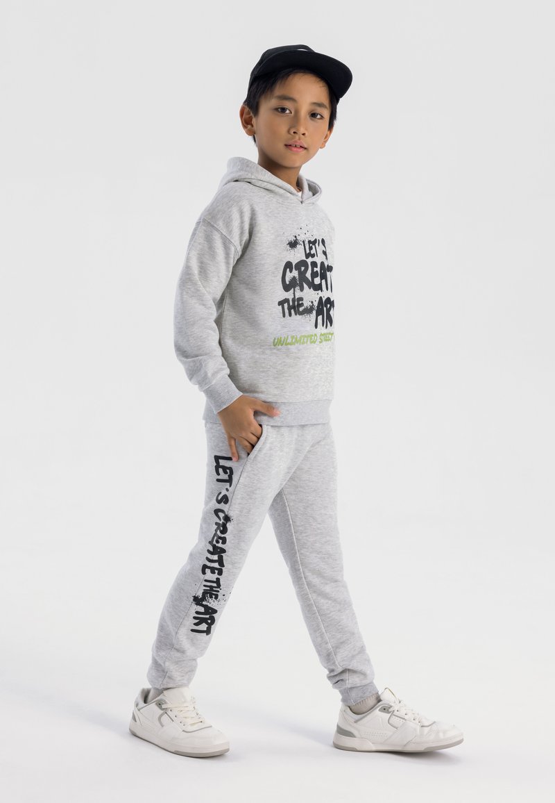 Grå bomullshoodie med grafiken "Låt oss skapa konsten" i svart och grönt. Matchande gråa sweatpants med sidofickor och liknande textdesign. Vita sneakers.