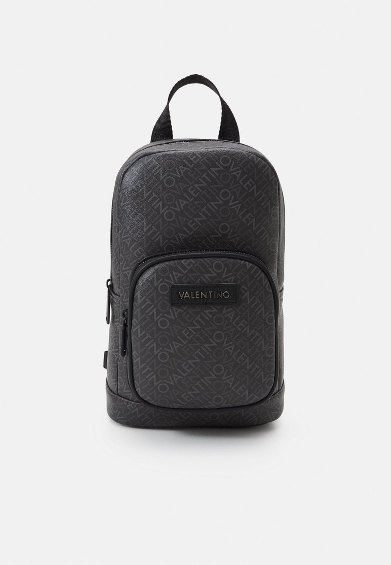 Schwarzer Valentino-Rucksack mit wiederholt auftretendem Logo-Muster, Frontreißverschlusstasche, Markenpatch und oberem Tragegriff vor weißem Hintergrund.