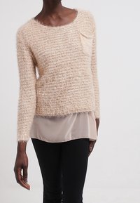 Ljust beige fluffig stickad tröja med rund halsringning, långa ärmar och en liten bröstficka, lagd över en genomskinlig, ljusfärgad topp.