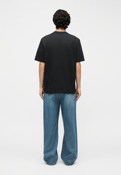 Άνδρας με σγουρά μαλλιά που φοράει απλό μαύρο κοντομάνικο μπλουζάκι και άνετο μπλε τζιν, όρθιος με την πλάτη προς τα εμάς σε λευκό φόντο.