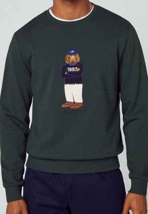 Ciemnozielona bluza z haftowanym psem w granatowej czapce, swetrze z napisem "1983", białych spodniach i brązowych butach, logo Hackett London poniżej.