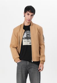 Tan spalvos bomber tipo striukė su elastinėmis rankovėmis ir juosmeniu, priekyje užtrauktukas. Po ja juodas grafiškas t-shirt'as. Pilkos džinsinės kelnės su subtiliu tekstūru.