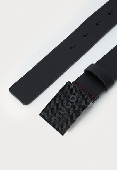 Bracelet de montre en caoutchouc noir avec une étiquette noire texturée, embossée du mot "HUGO". Il présente des coutures rouges et plusieurs trous d'ajustement sur toute sa longueur.