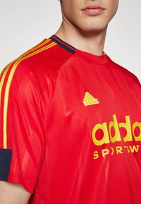 Červený sportovní dres s vertikálními pruhy, žlutými akcenty a modrým límcem. Výrazné logo "adidas" v žluté na hrudi.