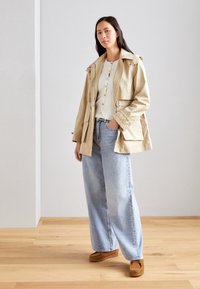 Beżowa, lekka kurtka zapinana na guziki, noszona na kremowym, prążkowanym topie oraz jasnoniebieskich, szerokich dżinsach. Brązowe zamszowe buty dopełniają look.