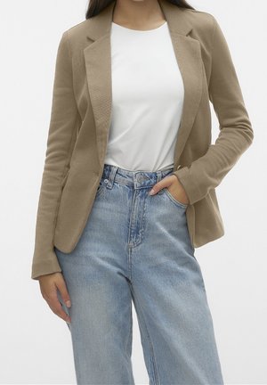 Woman wearing a beige blazer, white top, and light blue high-waisted jeans, with one hand in pocket and the other relaxed by side.